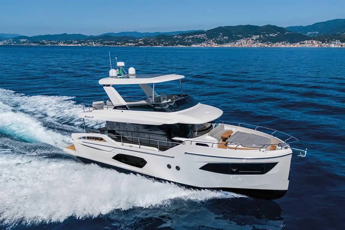 A sleek white motor yacht, Absolute 57, moves through blue waters with a scenic coastline in the background.